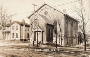 A Sepia Photo Shows A Brick Church With Tall Arched Windows Beside A Street, With Rail Tracks In The Foreground And Leafless Trees Around The Building.