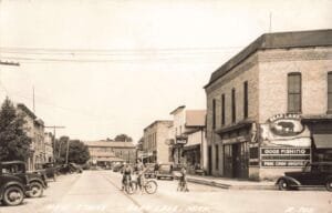 Black-And-White Street Scene With Vintage Cars Parked Along Both Sides, Three Kids On Bicycles In The Road, And A Corner Building With A Large “Bear Lake” Fishing And Camping Sign.