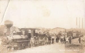 Sepia-Toned Photo Of A Steam Locomotive And Log Cars With A Large Group Of People Standing Beside The Train Near An Industrial Mill With Tall Smokestacks.