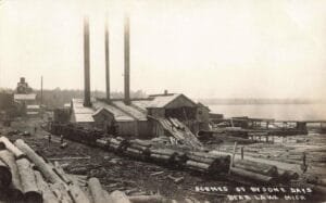 Black-And-White Photo Of A Shoreline Sawmill With Three Smokestacks, Log Piles And A Long Ramp Or Chute Leading Toward The Water At Bear Lake.