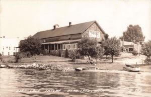 Black-And-White Photo Of The Bear Lake Tavern On A Shoreline With A Porch, Small Dock And Rowboats On The Water In The Foreground.