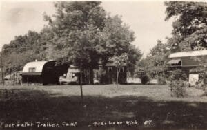 Black-And-White Photo Of Two Travel Trailers And An Older Car Parked On A Grassy Campground Beneath Large Trees.