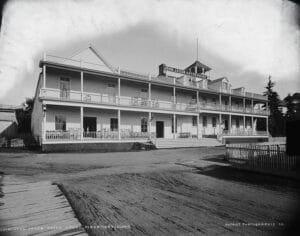 Black-And-White View Of A Long, Two-Story Hotel With Wide Porches And A Rooftop Sign Reading “John Jacob Astor House,” Fronting A Dirt Street And Picket Fence.