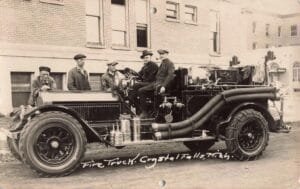 Black-And-White Photo Of Five Men Posing With An Early Open-Cab Fire Truck With Large Hoses And Tire Chains, Parked Beside A Brick Building.
