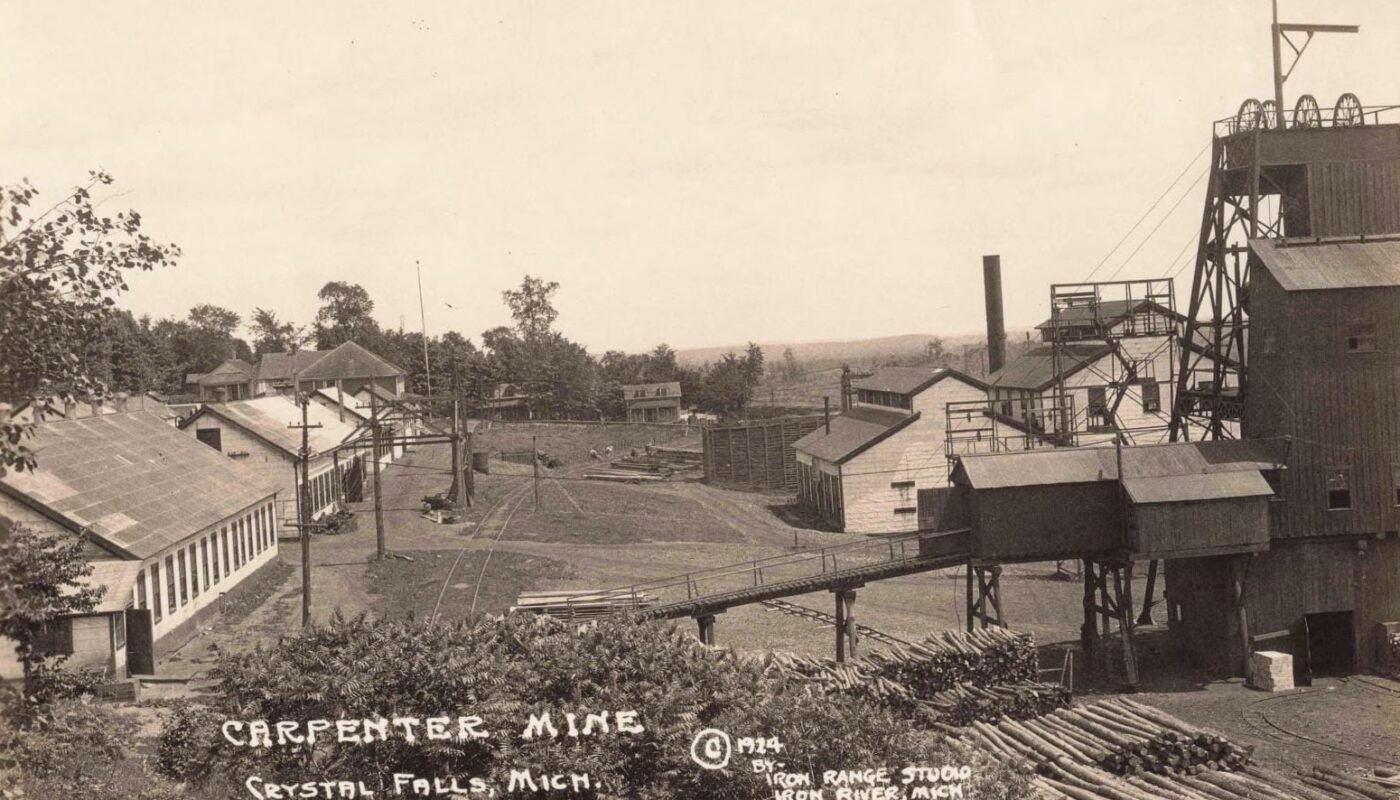 Black-And-White View Of An Iron Mine Complex With A Tall Headframe, Industrial Buildings, Rail Tracks, And Piles Of Cut Logs In The Foreground.