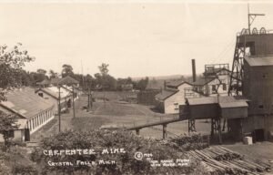 Black-And-White View Of An Iron Mine Complex With A Tall Headframe, Industrial Buildings, Rail Tracks, And Piles Of Cut Logs In The Foreground.