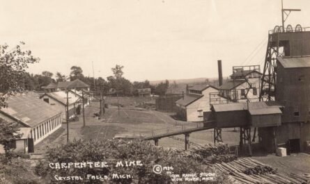 Black-And-White View Of An Iron Mine Complex With A Tall Headframe, Industrial Buildings, Rail Tracks, And Piles Of Cut Logs In The Foreground.