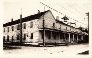 Black-And-White Photo Of A Long, Two-Story Wooden Building With A Covered Porch And A Sign Reading “John Jacob Astor House” Along A Street With Utility Poles.