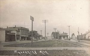 Railroad Tracks Curve Past A Depot And Freight Cars, With Storefronts, An Early Automobile And Utility Poles Along The Street At Left.