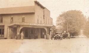 Historic Photograph Of The Clarksville Oil And Gas Company In Clarksville, Michigan, With Two Automobiles, Gasoline Pumps, Tire Advertisements And Two Men Outside The Building.