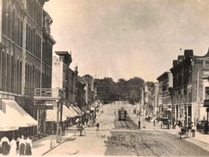 Historic View Of Main Street In Niles, Michigan, With The Michigan Inn Sign, Pedestrians, Downtown Storefronts And An Electric Railway Car Running Along Tracks In The Street.