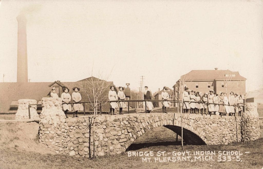 Children Stand Along A Railing Above A Stone Bridge, With School Buildings In The Background.