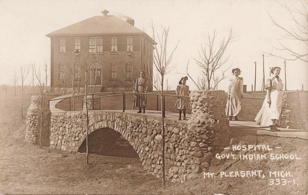 Four People Stand On A Stone Bridge Beside A Two-Story Brick Building, With “Hospital — Gov’t. Indian School — Mt. Pleasant, Mich.” Written On The Photograph.