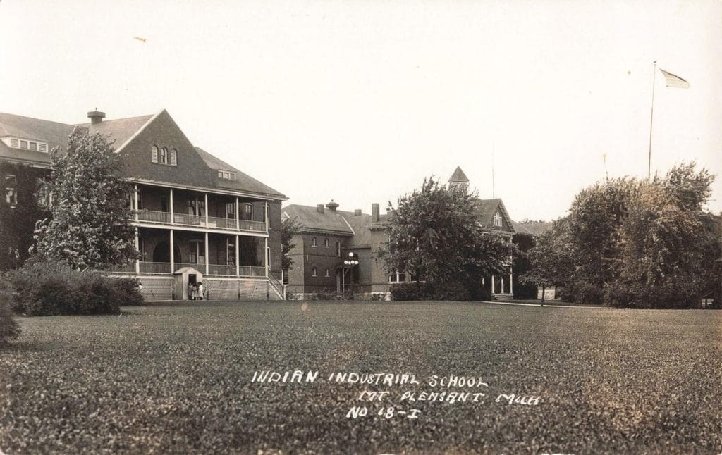 Brick Buildings Stand Behind A Wide Lawn, With Two Small Figures Near A Doorway At Left And A Flagpole At Right.