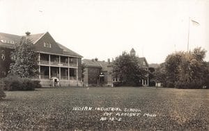 Brick Buildings Stand Behind A Wide Lawn, With Two Small Figures Near A Doorway At Left And A Flagpole At Right.