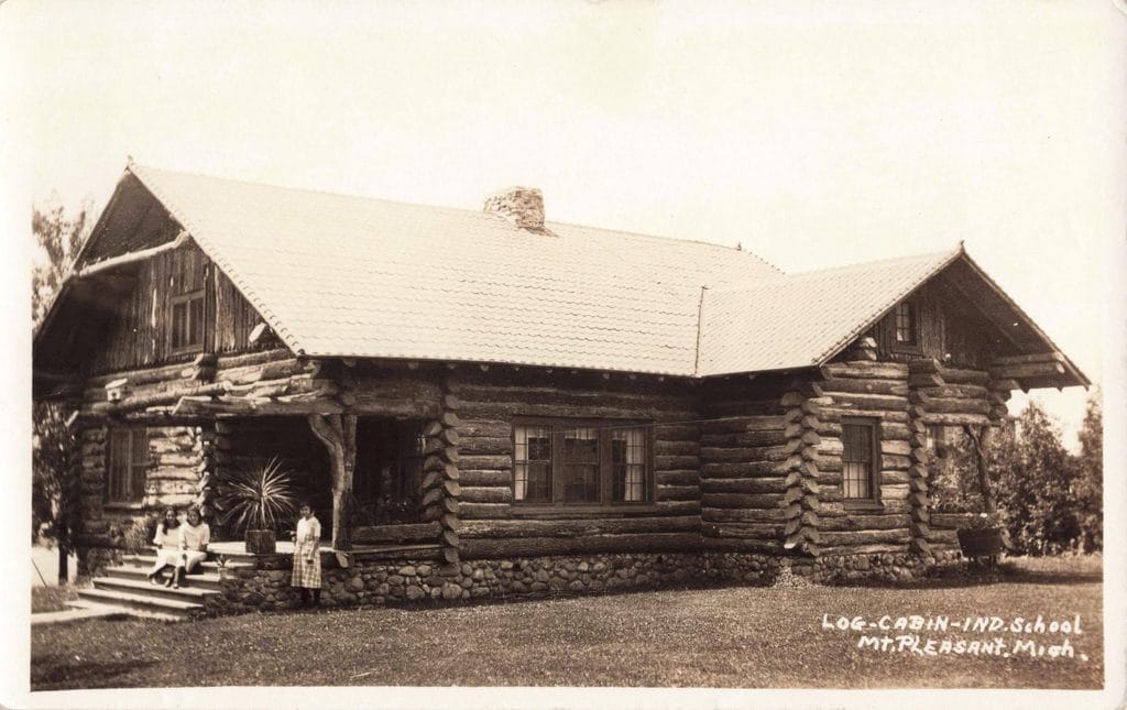 Two Young People Sit On Porch Steps And A Third Stands Beside A Large Log Building With A Stone Foundation, Gabled Roof And Stone Chimney.