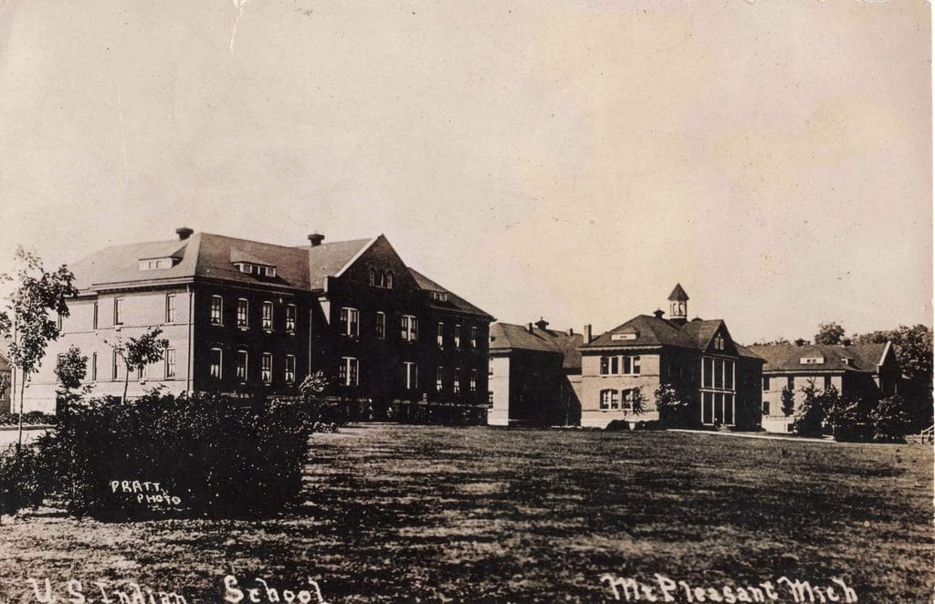 Mount Pleasant Indian Boarding School - Several Multistory Brick Buildings With Pitched Roofs Stand Beyond A Lawn And Shrubs, With A Small Tower Above The Building Near The Center-Right.