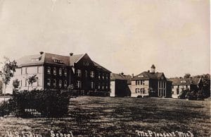 Several Multistory Brick Buildings With Pitched Roofs Stand Beyond A Lawn And Shrubs, With A Small Tower Above The Building Near The Center-Right.