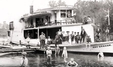 The May Graham Riverboat Is Tied To A Dock As Passengers Stand Aboard And Children Wade In The River.