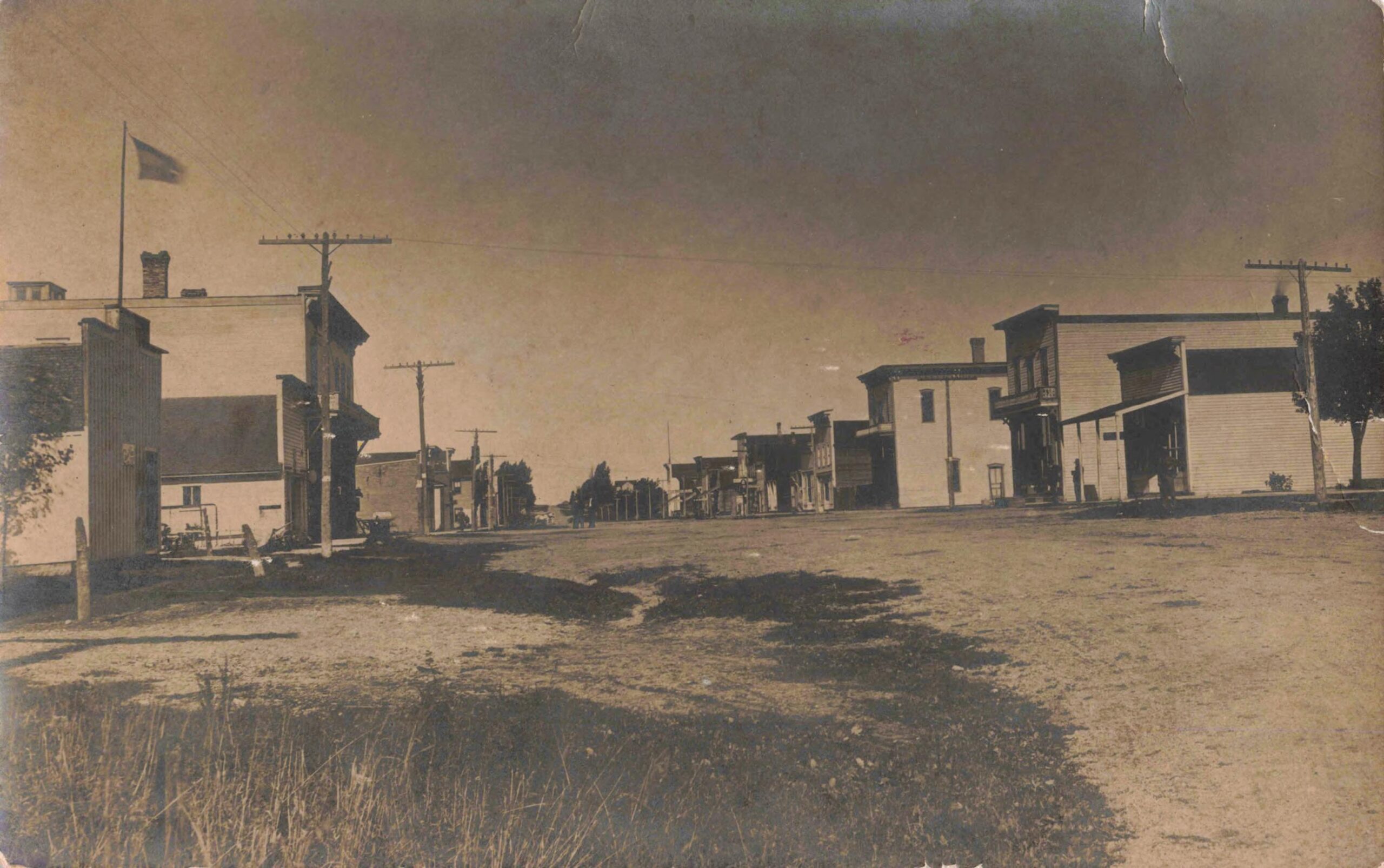 Wide View Of Mecosta's Unpaved Commercial Street With Wood-Frame Stores, Utility Poles And Pedestrians In The Early 1900S